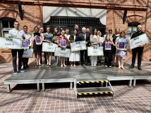 Grupowe zdjęcie laureatów piątej edycji konkursu GOZ – Lider Małopolski. Przed ceglanym budynkiem stoi kilkanaście osób reprezentujących nagrodzone firmy, szkoły i organizacje pozarządowe. Uczestnicy trzymają dyplomy, statuetki oraz duże czeki z nagrodami o wartości od 2 000 do 5 000 zł. W dolnej części grafiki znajduje się fioletowy baner z napisem „Laureaci piątej edycji konkursu” oraz logo konkursu.